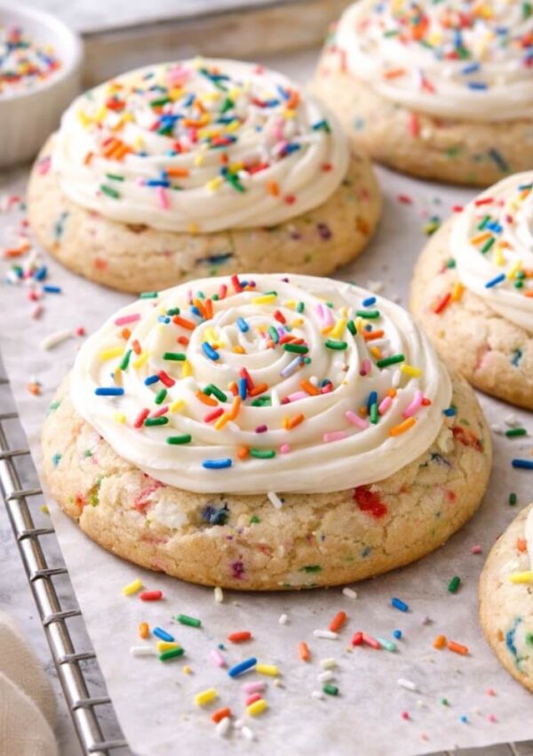 A close-up of large, round sugar cookies with colorful sprinkles inside, topped with swirls of white frosting and more rainbow sprinkles, displayed on parchment paper over a cooling rack.