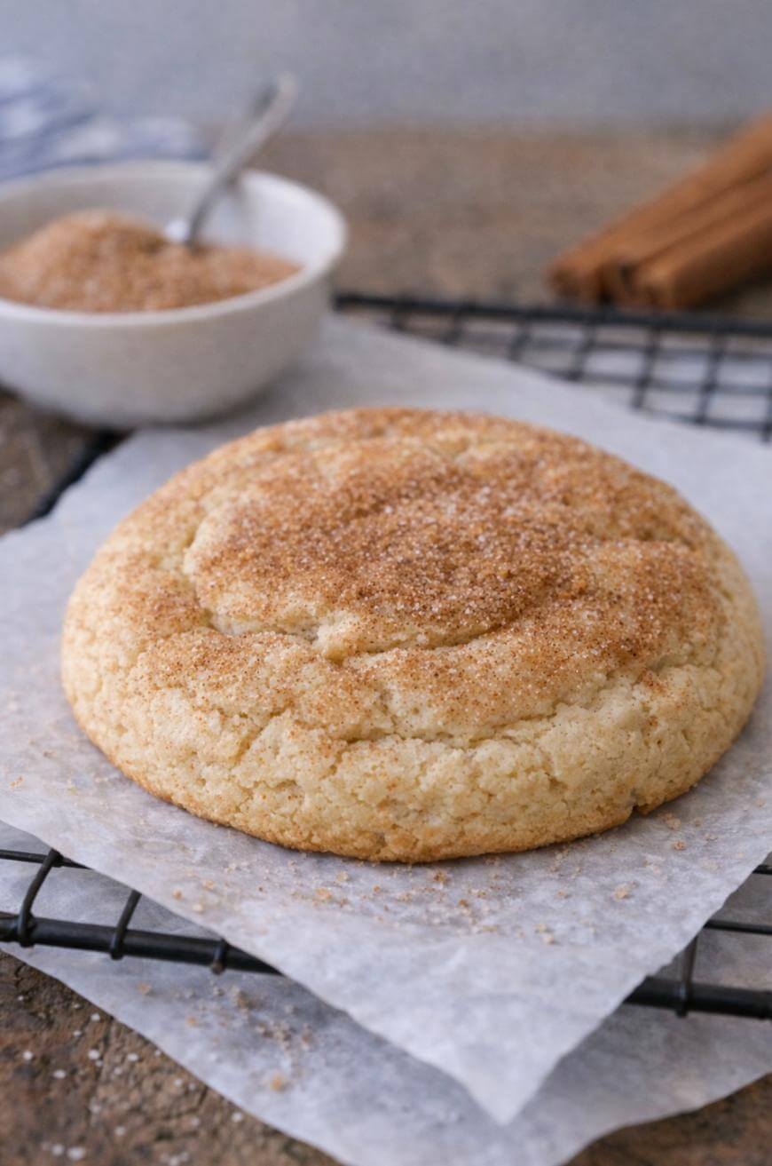 A large, round snickerdoodle cookie dusted with cinnamon sugar sits on parchment paper atop a cooling rack, with a bowl of cinnamon sugar and cinnamon sticks in the background.
