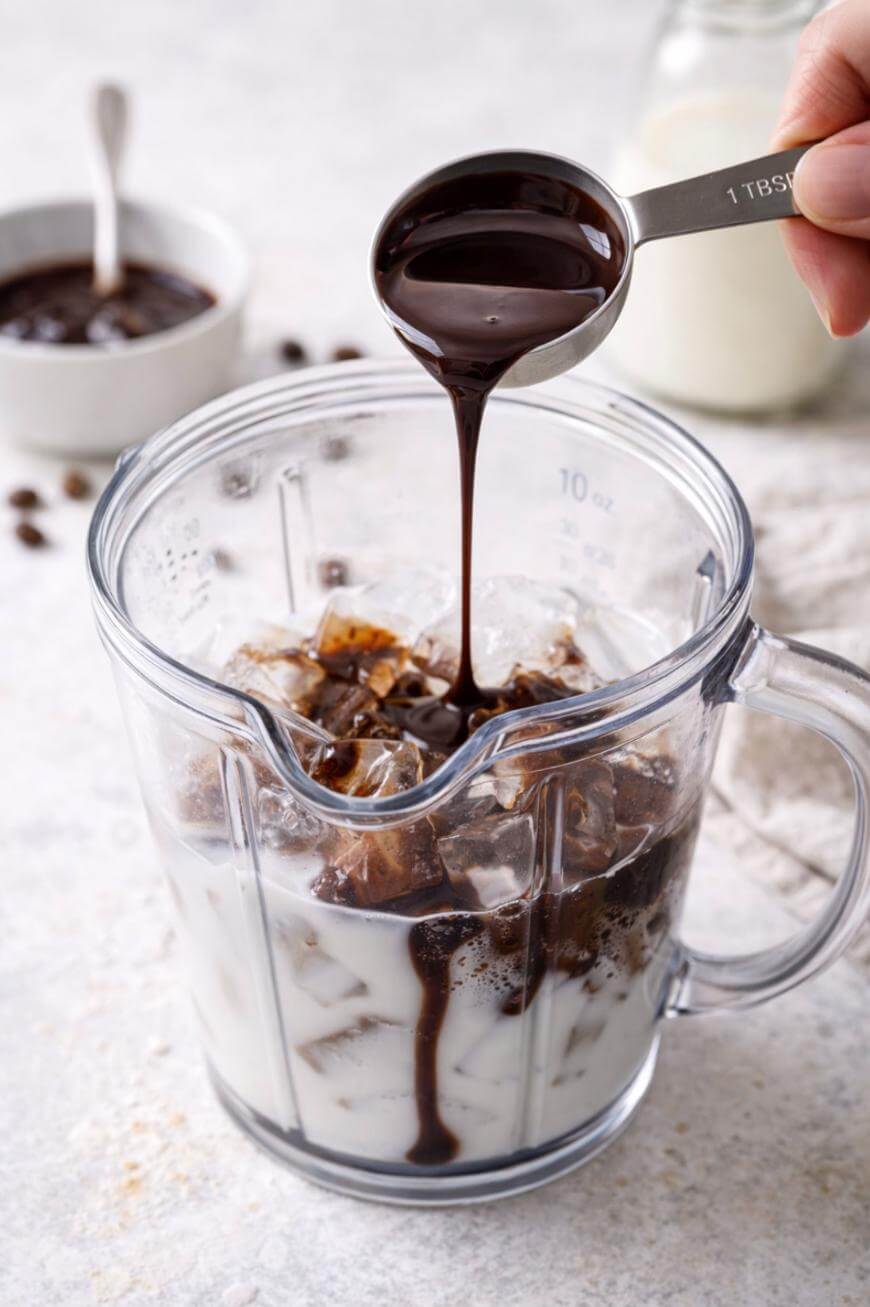 A tablespoon of chocolate syrup is being poured into a blender filled with ice, milk, and coffee cubes. A blurred jar and bowl are visible in the background.
