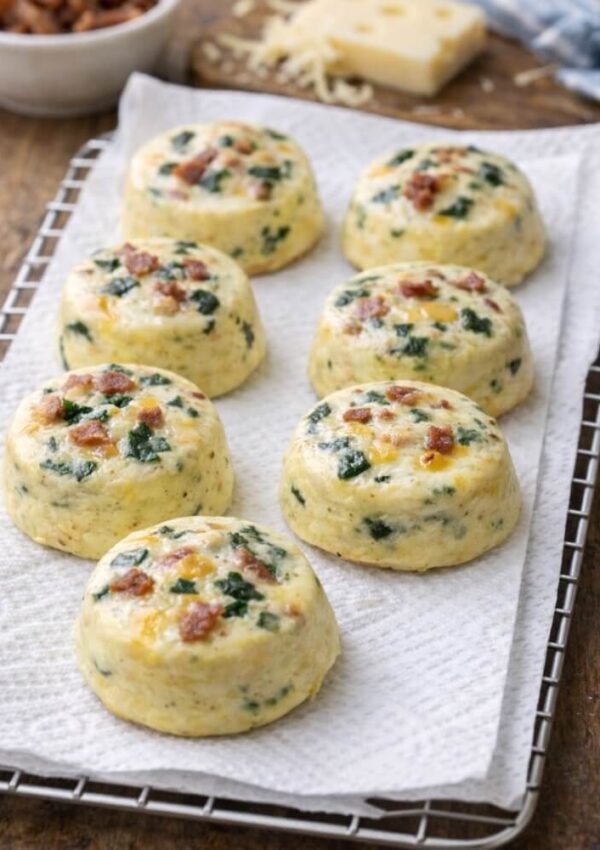 Six baked egg bites with spinach and bacon pieces are arranged on a paper towel over a cooling rack. A bowl of ingredients and a block of cheese are visible in the blurred background.
