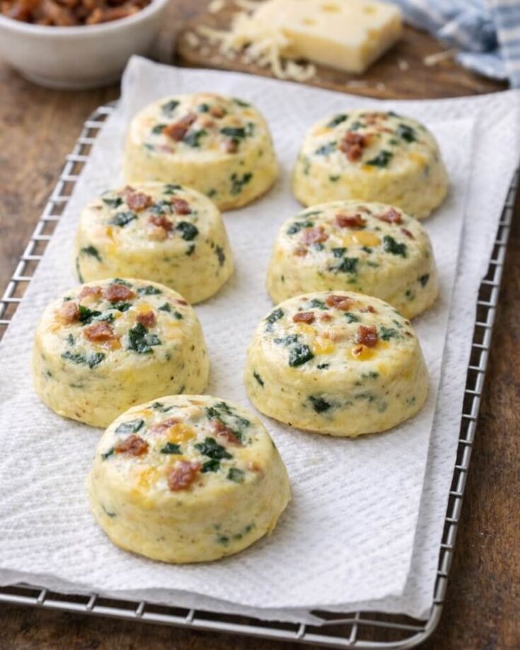 Six baked egg bites with spinach and bacon pieces are arranged on a paper towel over a cooling rack. A bowl of ingredients and a block of cheese are visible in the blurred background.