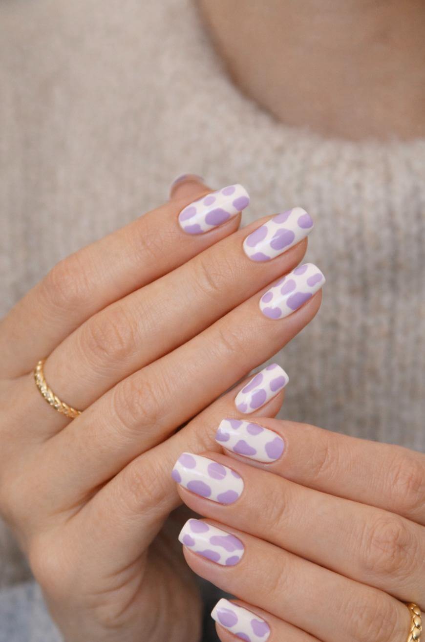 Close-up of hands with long nails painted white and decorated with irregular lavender and purple spots. The person wears a gold ring and a beige knit sweater.