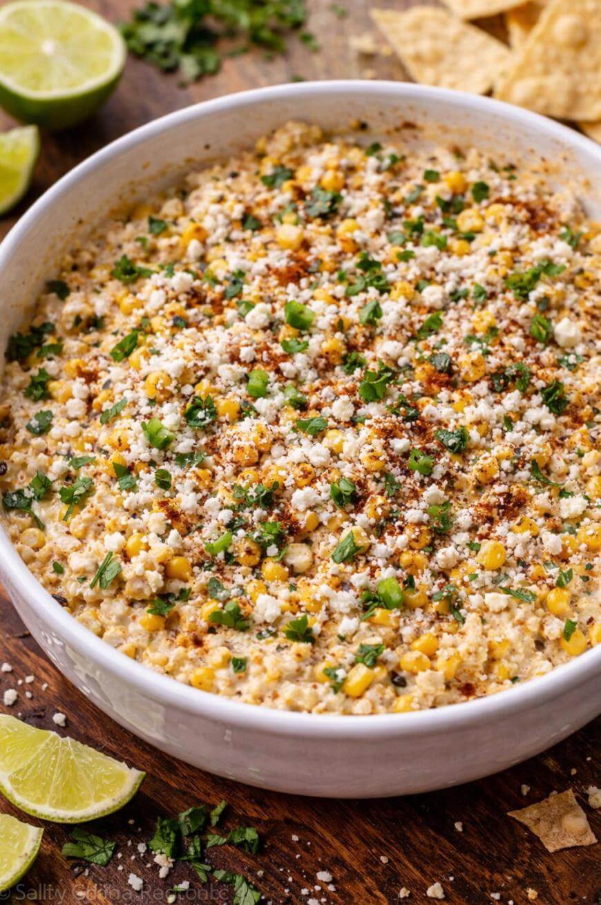 A bowl of Mexican street corn dip garnished with crumbled cheese, chopped cilantro, green onions, and spices. Lime wedges and tortilla chips are visible on the wooden surface beside the bowl.