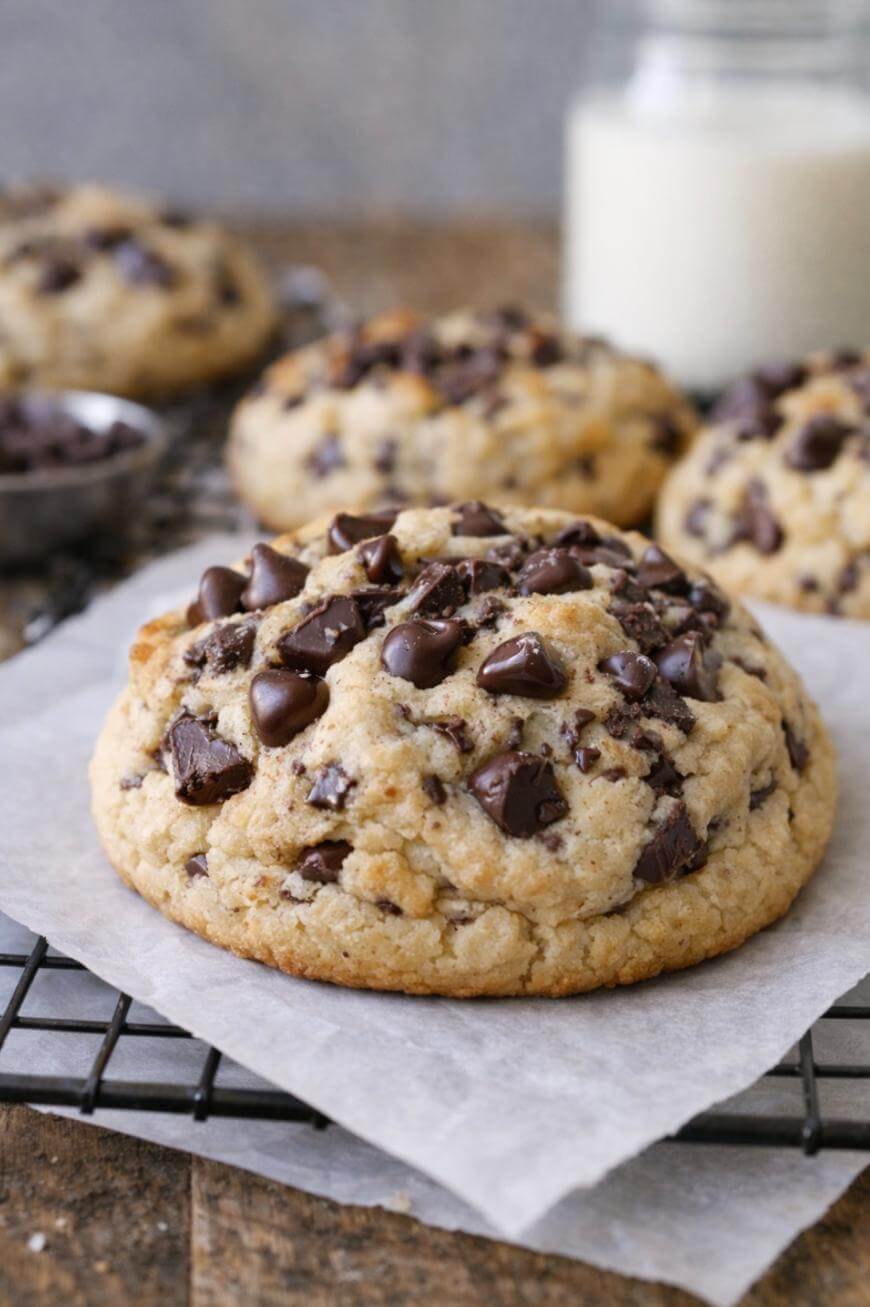 A thick, homemade chocolate chip cookie sits on parchment paper on a cooling rack, with more cookies and a jar of milk blurred in the background.