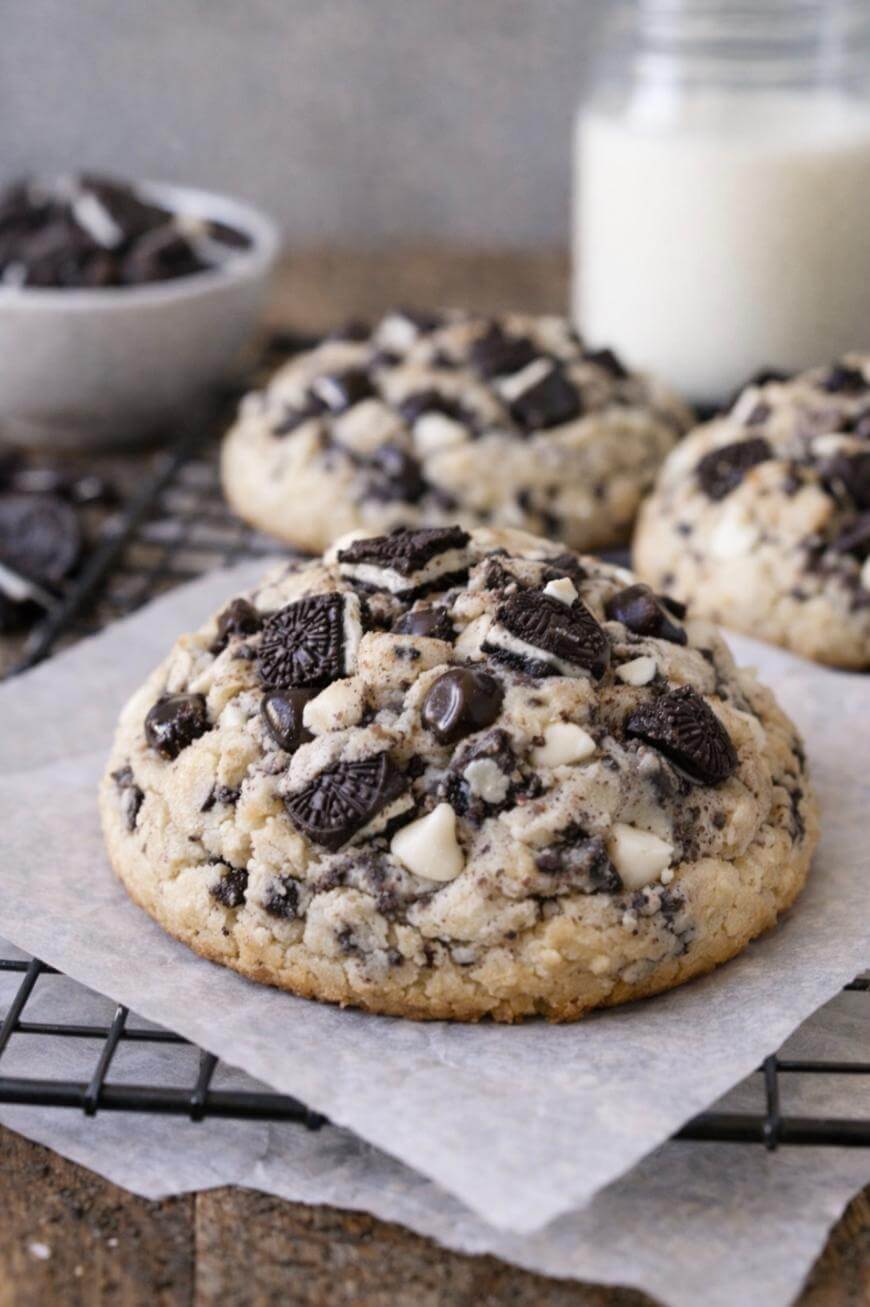 Three large cookies with chocolate chips and Oreo pieces are on parchment paper atop a cooling rack. In the background, there is a bowl of cookie pieces and a jar of milk.
