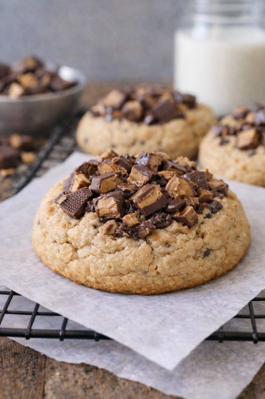A thick, chewy cookie topped with chunks of chocolate and peanut butter candies sits on parchment paper on a cooling rack, with more cookies and a glass of milk in the background.