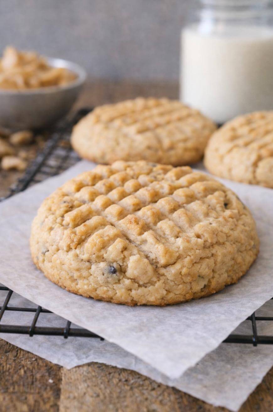 A close-up of a peanut butter cookie with a crisscross pattern on top, resting on parchment paper on a cooling rack. More cookies, a bowl of peanuts, and a jar of milk are blurred in the background.