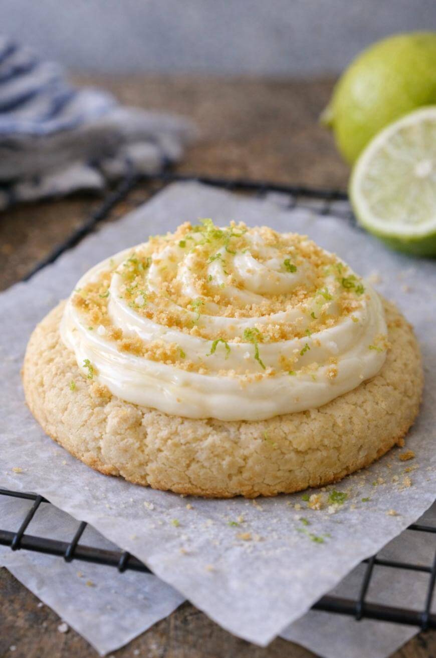 A large cookie topped with swirled cream cheese frosting, crushed graham crackers, and lime zest sits on parchment paper with limes in the background.