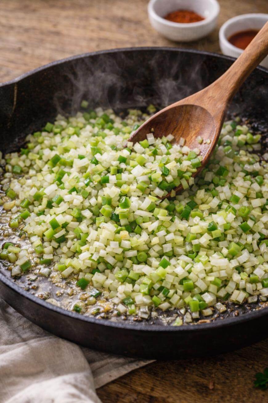 Diced green bell peppers and onions being sautéed in a cast iron skillet with a wooden spoon, with steam rising and spices in small bowls in the background.