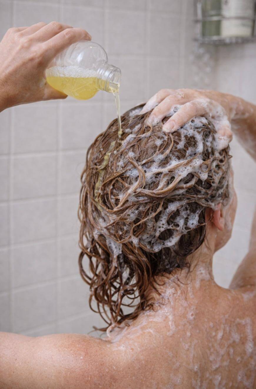A person in a shower pours liquid shampoo onto their wet, lathered hair, which is covered in thick foam. The tiled shower wall and a metal shelf with bottles are visible in the background.