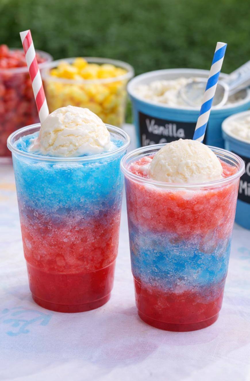 Two clear cups filled with red and blue shaved ice, each topped with a scoop of vanilla ice cream and a striped straw. Containers of toppings and ice cream are visible in the blurred background.