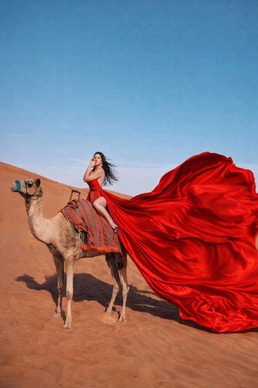 A woman in a flowing red dress sits on a camel in a sandy desert. The fabric of her dress billows dramatically in the wind, creating a striking contrast against the blue sky and orange dunes.