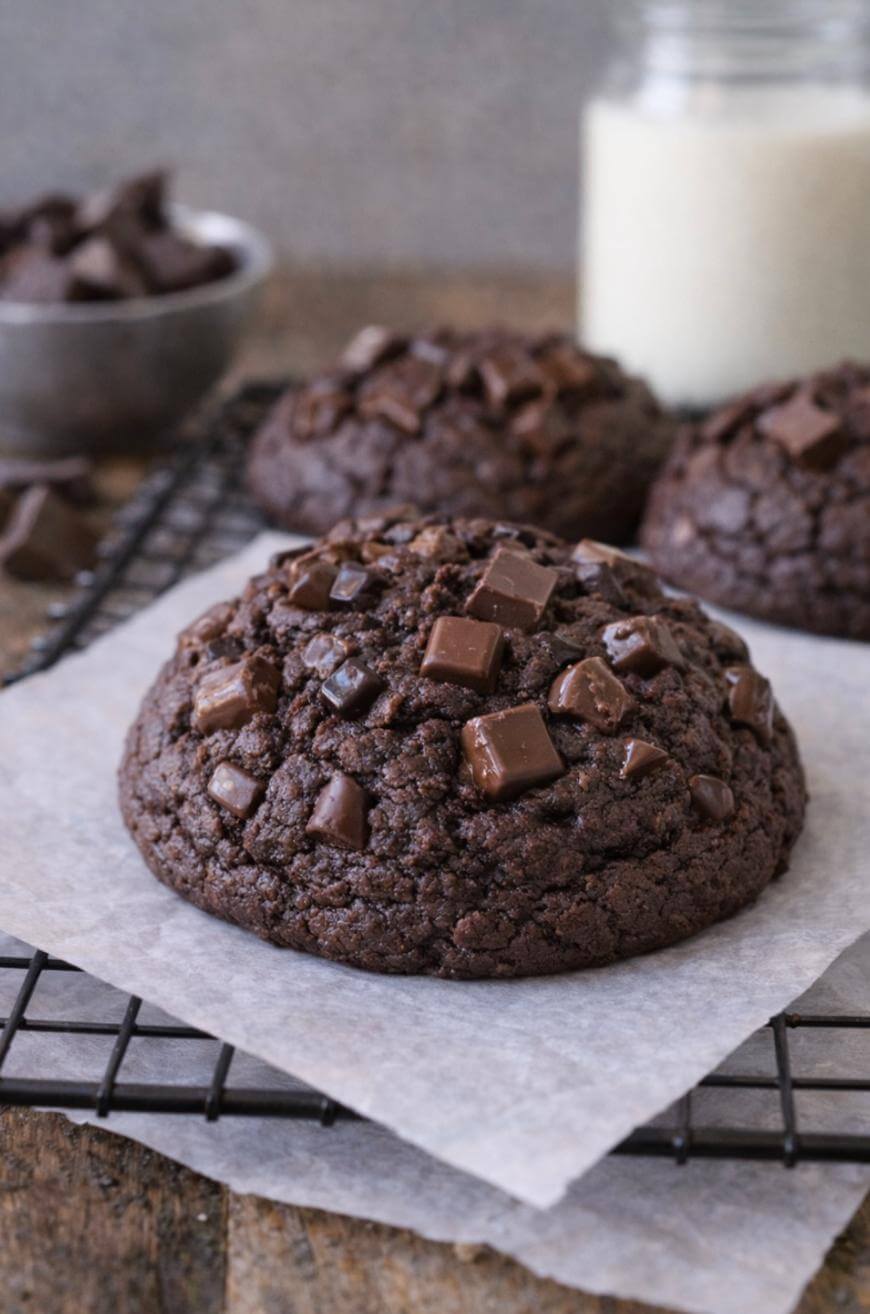 A large, thick double chocolate cookie with chunky chocolate pieces sits on parchment paper atop a cooling rack, with more cookies and a glass of milk in the background.