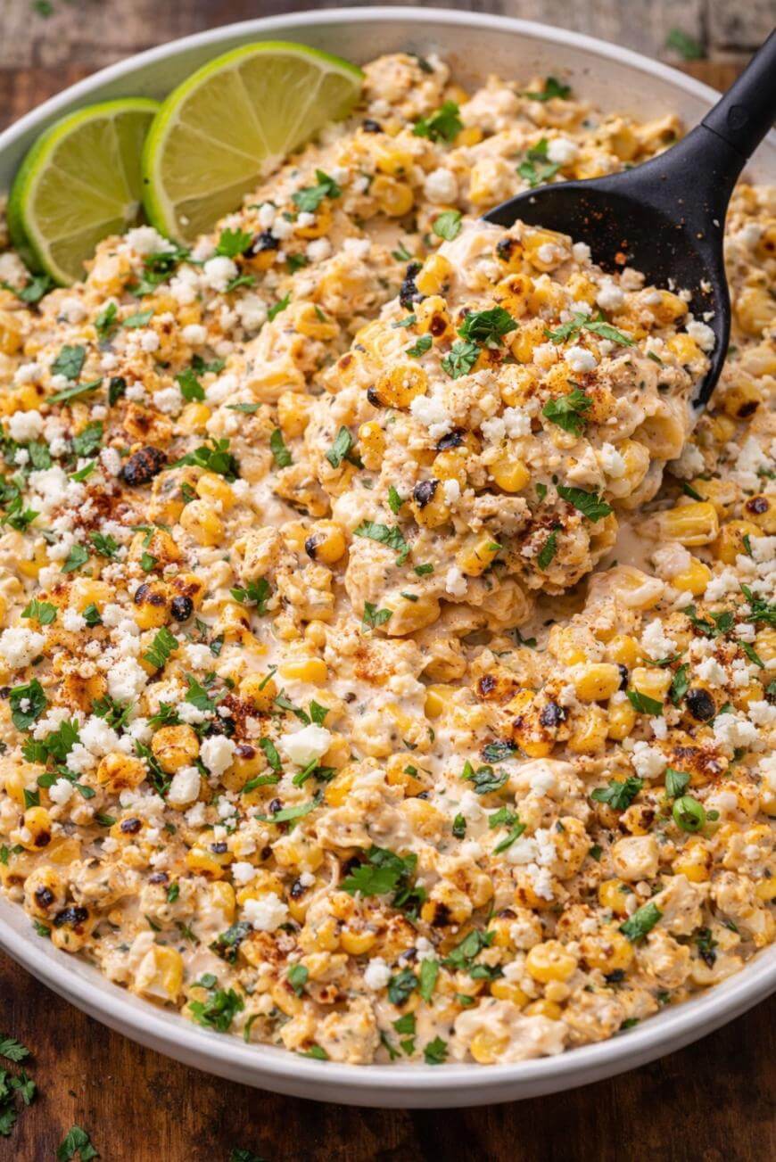 A bowl of creamy Mexican street corn salad topped with crumbled cheese, chopped cilantro, and chili powder, with lime wedges on the side and a black spoon scooping some of the salad.