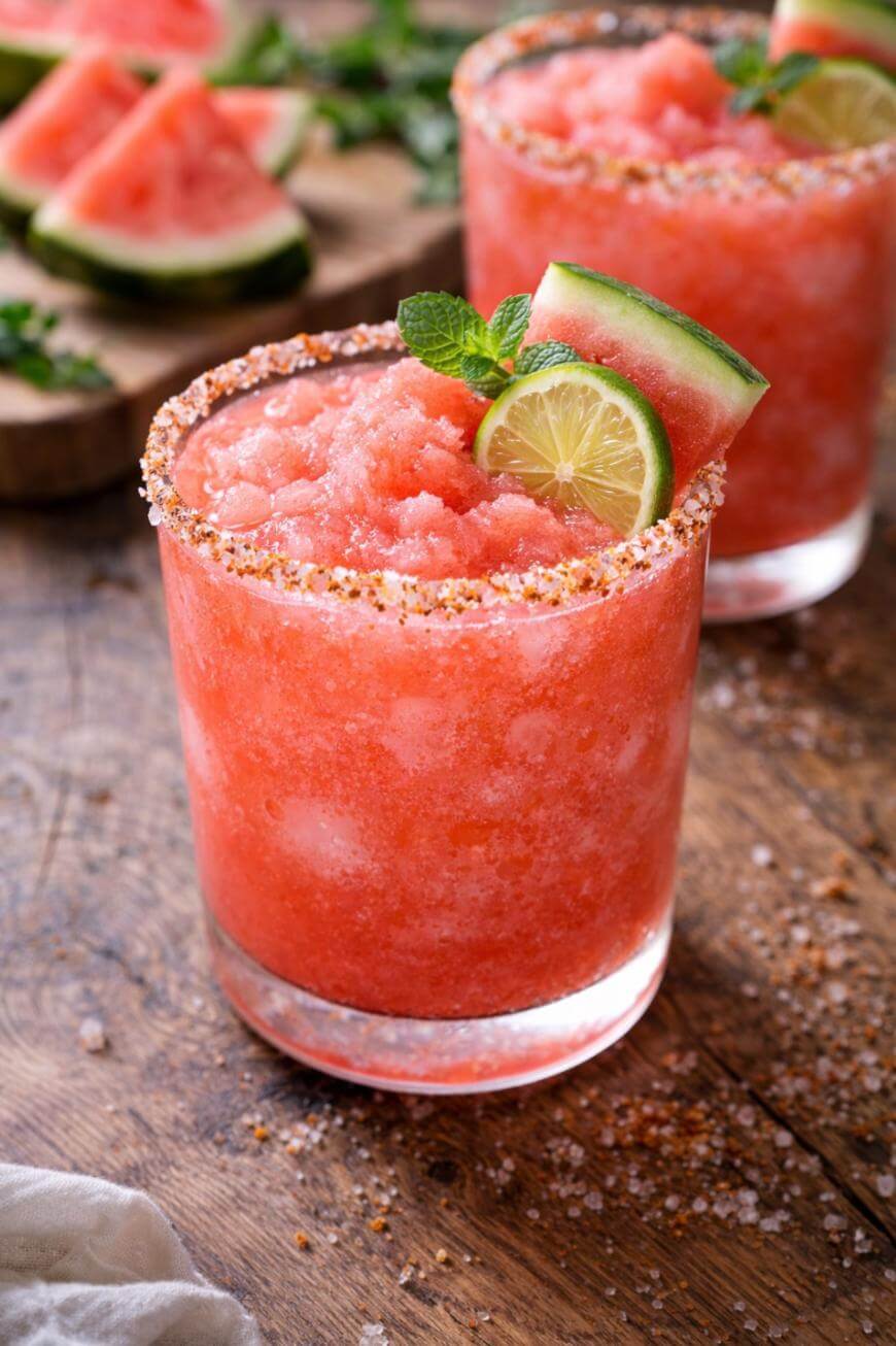 Two glasses of frozen watermelon drink with salt and chili powder rims, garnished with lime slices, watermelon wedges, and mint, sit on a rustic wooden table. More watermelon slices are in the background.