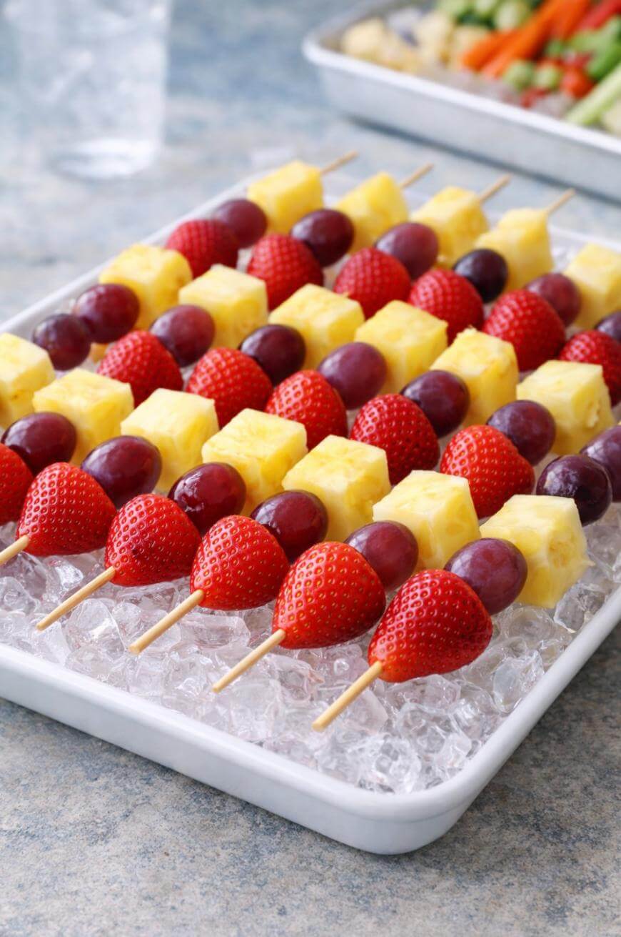 A tray of fruit skewers with strawberries, grapes, and pineapple chunks arranged in neat rows on a bed of ice. Another tray with vegetables is blurred in the background.