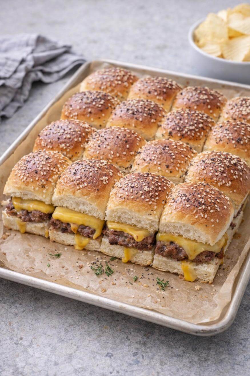 A tray of freshly baked cheeseburger sliders topped with sesame seeds, filled with melted cheese and beef patties, sits on parchment paper. A bowl of potato chips and a folded napkin are in the background.
