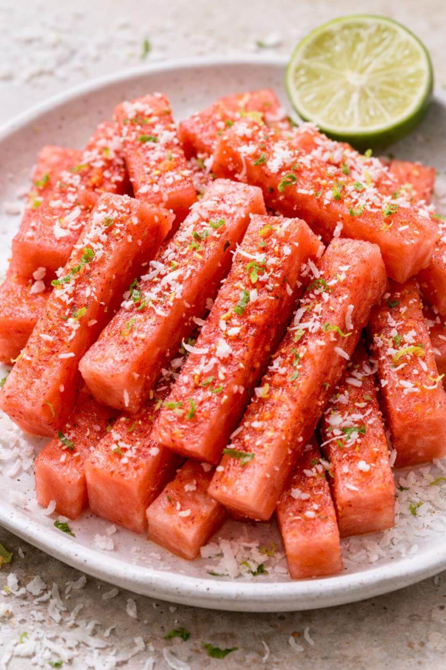 A plate of watermelon sticks topped with shredded coconut, chili powder, lime zest, and a halved lime in the background. The watermelon is arranged neatly on a white plate.