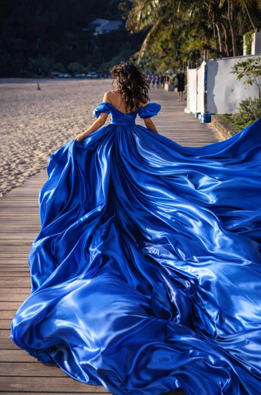 A woman with curly hair walks on a wooden boardwalk beside a beach, wearing a dramatic, flowing royal blue gown that spreads out behind her, catching the sunlight. Palm trees and greenery line the background.