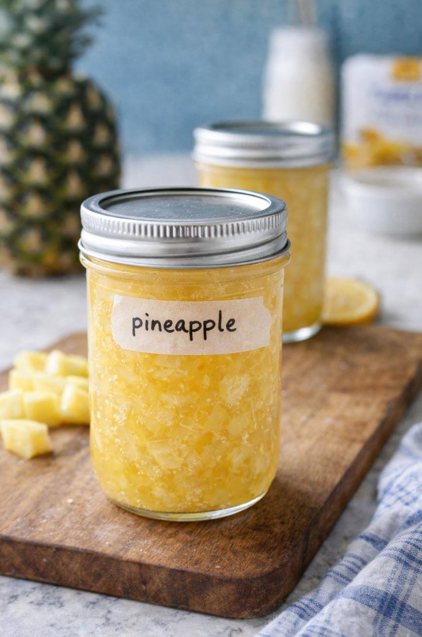 A glass jar labeled pineapple filled with crushed pineapple sits on a wooden board, with another jar and pineapple chunks in the background. A whole pineapple is also partially visible.