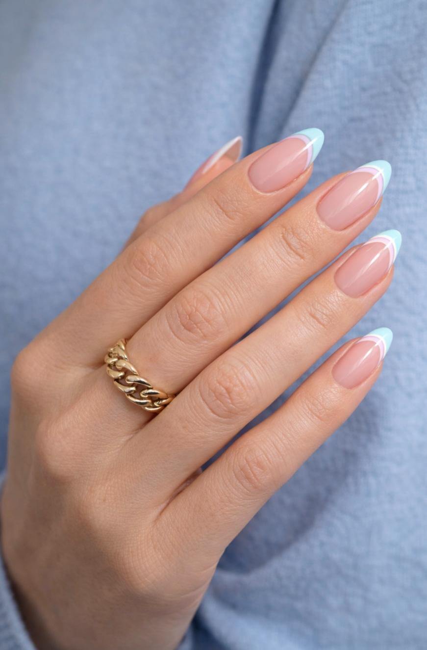 A hand with almond-shaped nails featuring a French manicure with light blue tips. The person wears a chunky gold chain ring and a pale blue long-sleeve top.