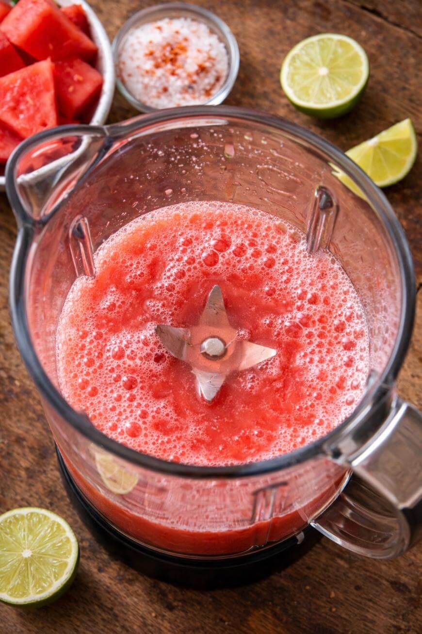 A blender filled with frothy, blended watermelon sits on a wooden surface, surrounded by lime halves, a bowl of salt with chili powder, and a dish of watermelon chunks.