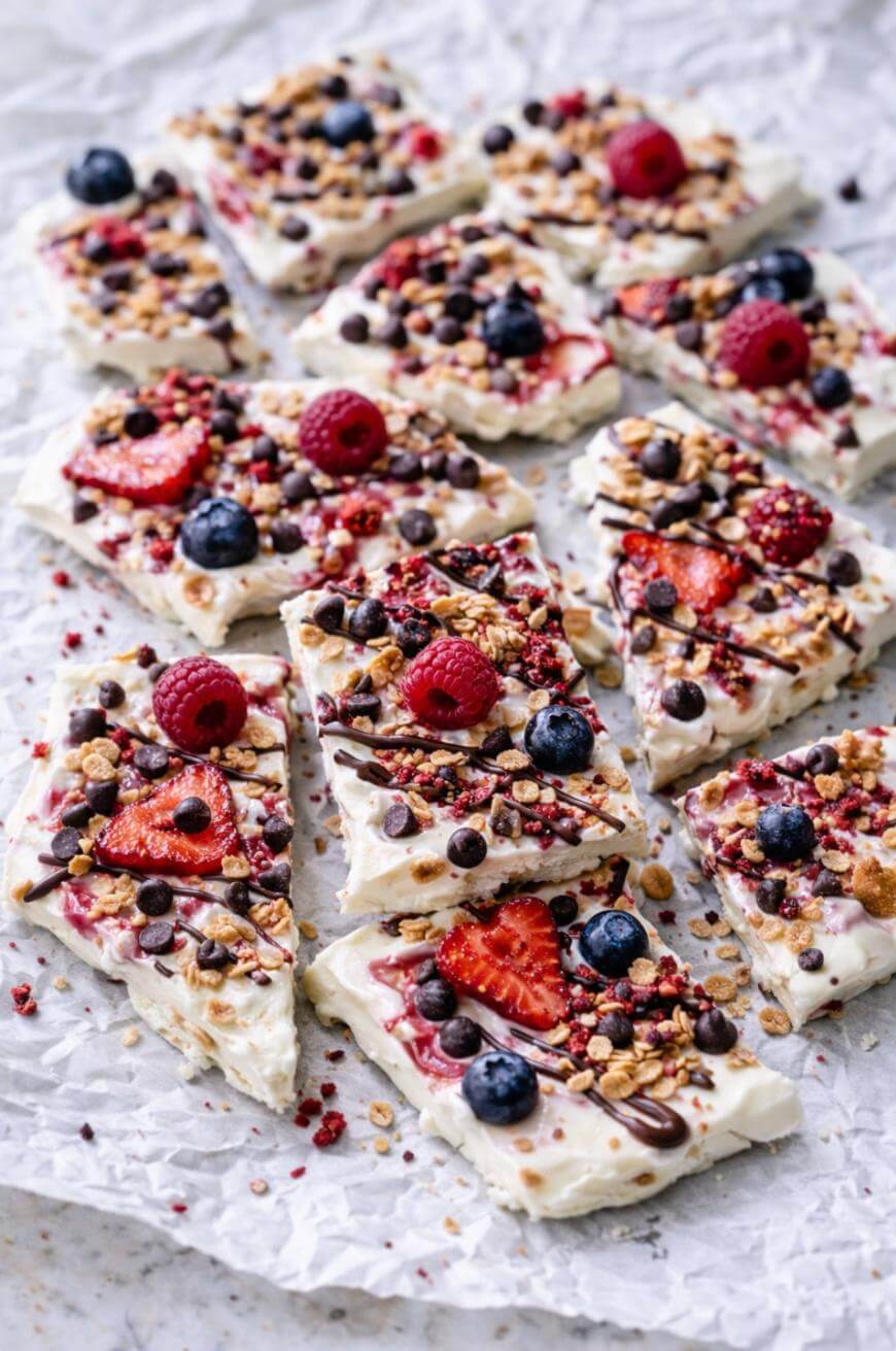 Sliced yogurt bark topped with fresh strawberries, blueberries, raspberries, granola, and chocolate chips, displayed on crumpled parchment paper.