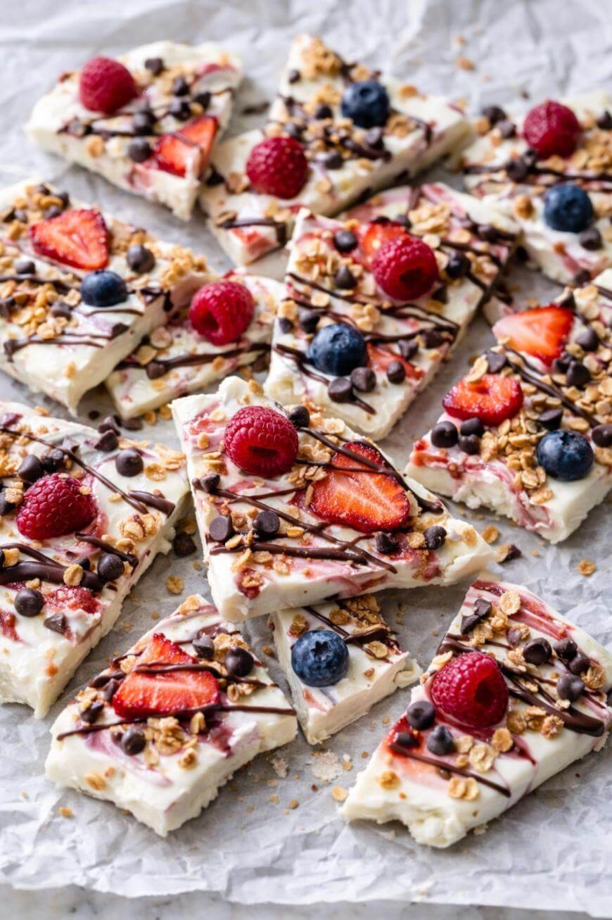 Several pieces of yogurt bark topped with sliced strawberries, raspberries, blueberries, granola, and a drizzle of chocolate, arranged on crumpled parchment paper.
