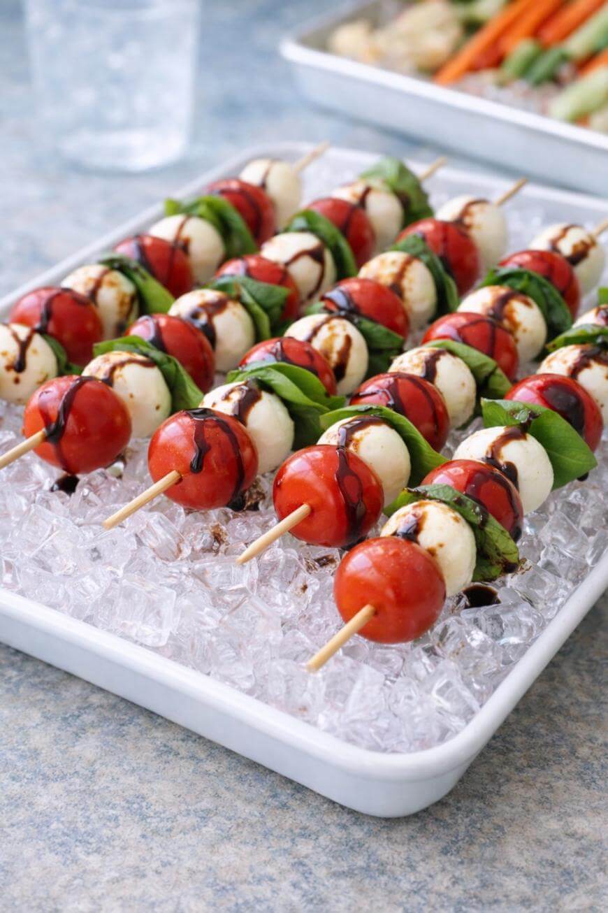 Skewers with cherry tomatoes, mozzarella balls, and basil leaves drizzled with balsamic glaze are arranged on a tray of ice, ready to serve as a fresh appetizer.