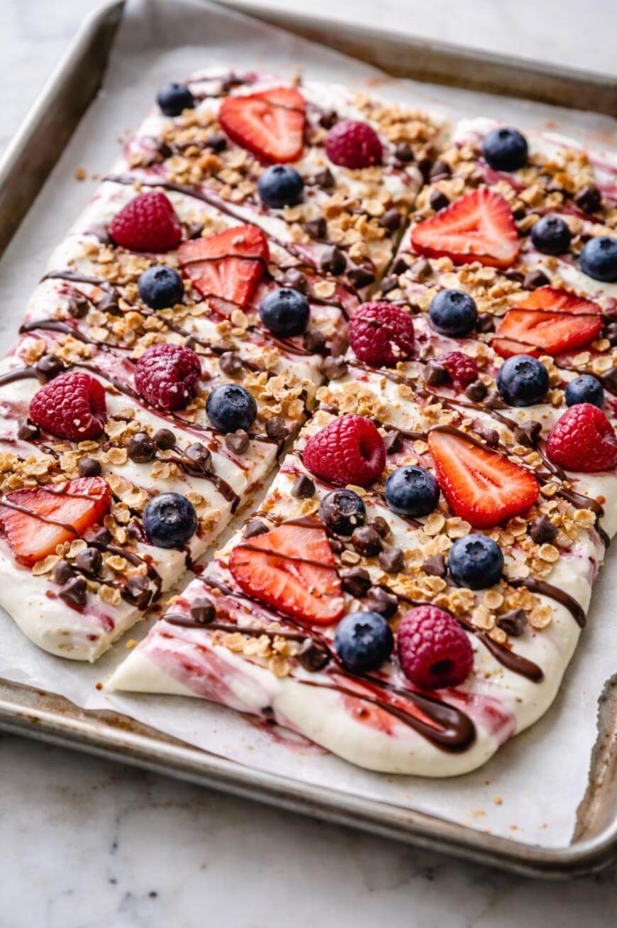 A rectangular dessert pizza topped with sliced strawberries, blueberries, raspberries, granola, and chocolate drizzle, sliced into six pieces on a baking sheet lined with parchment paper.