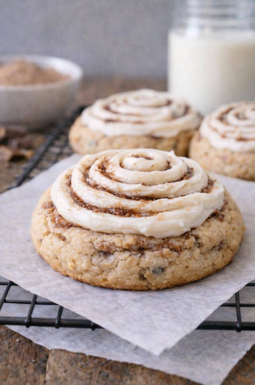 A close-up of a cookie topped with swirled icing, resembling a cinnamon roll, sitting on parchment paper on a cooling rack. More similar cookies and a glass of milk are blurred in the background.