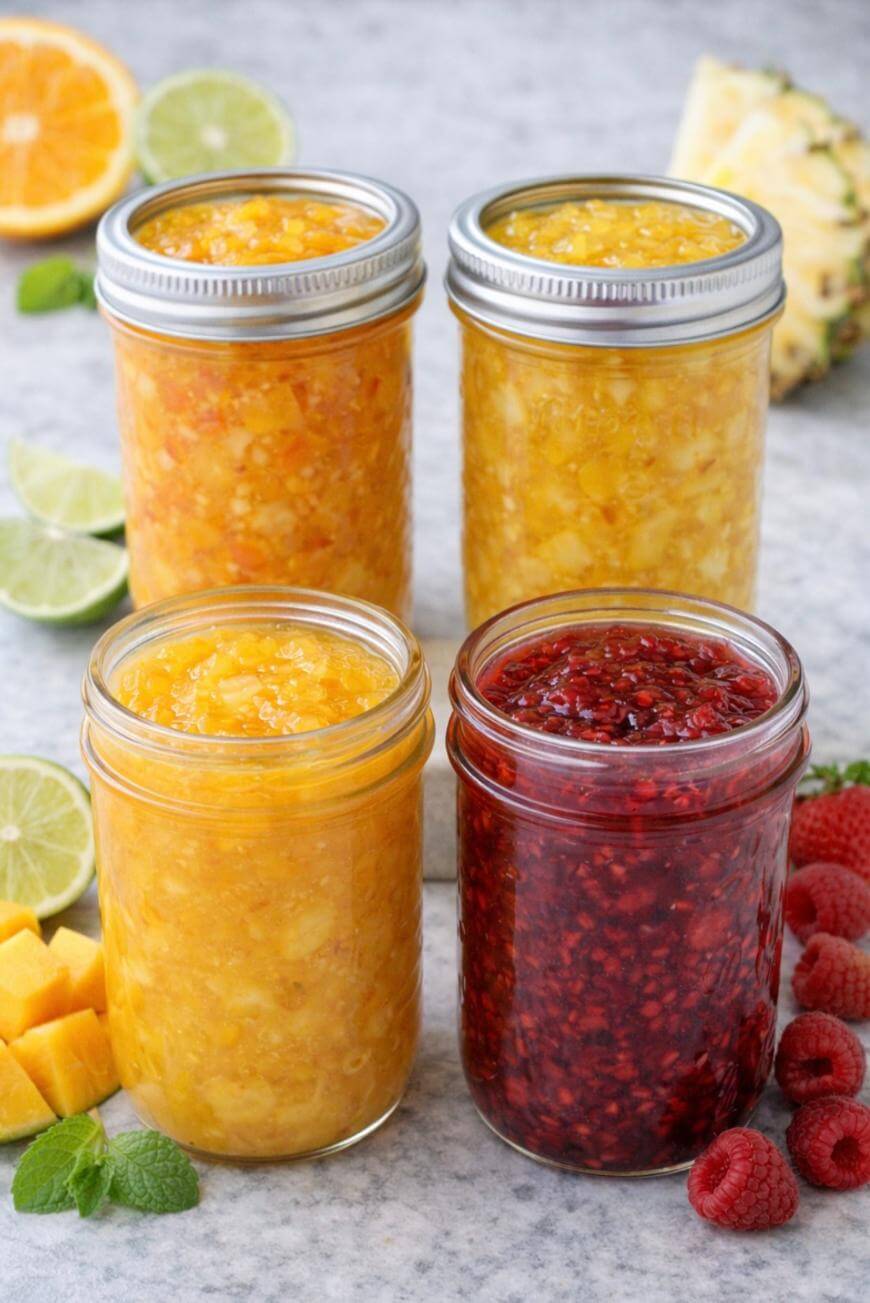 Four jars of homemade jam in different colors—yellow, orange, and red—are arranged on a gray surface with fresh fruit such as raspberries, mango, lime, orange, and pineapple around them.