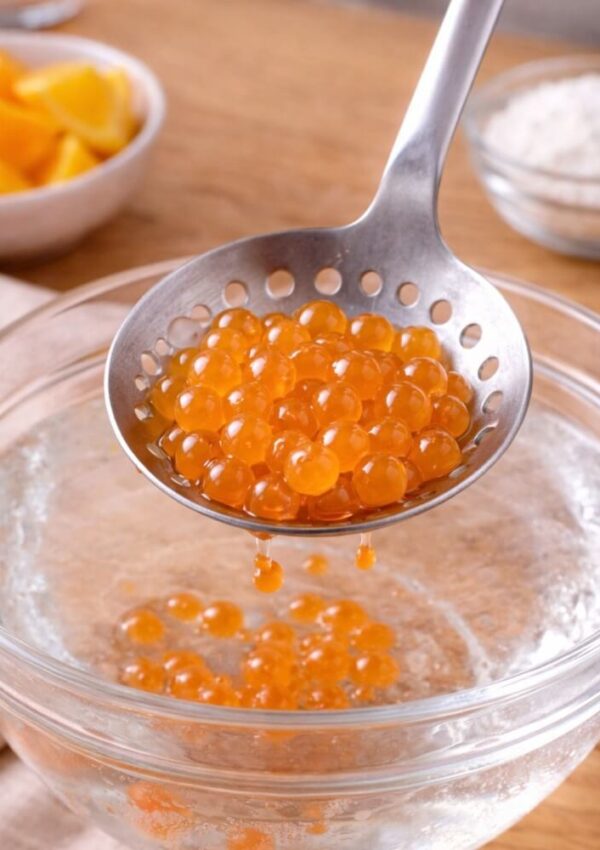 A slotted spoon holds bright orange spheres over a bowl of clear liquid, with lemon wedges and a bowl of white powder in the background on a wooden surface.