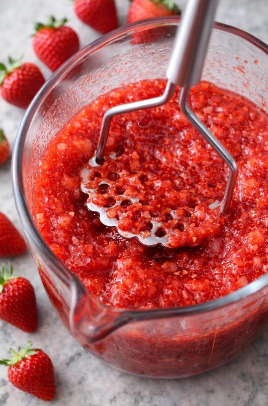 A glass bowl filled with mashed strawberries, being pressed with a metal potato masher. Fresh whole strawberries are scattered on the gray countertop nearby.