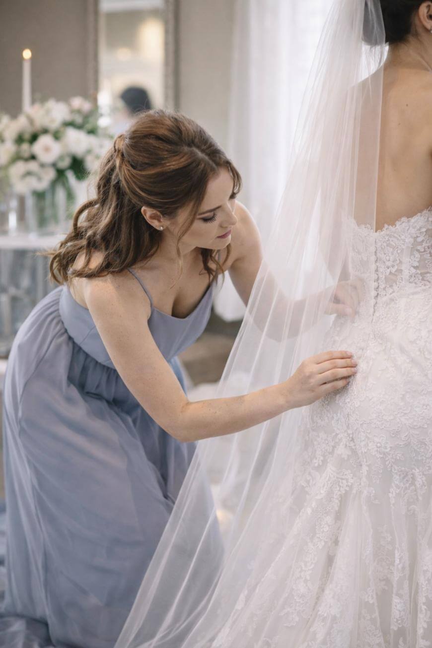 A bridesmaid in a lavender dress adjusts the back of a bride’s lace wedding gown and veil in a softly lit room decorated with white flowers.