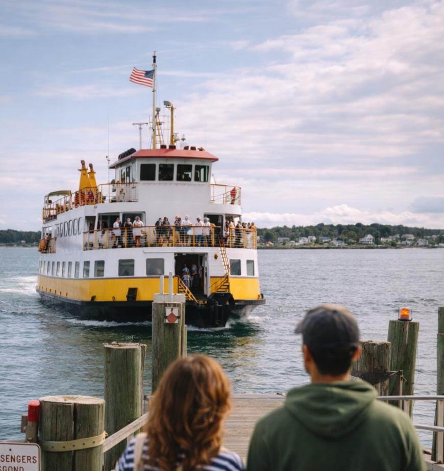 A yellow and white ferry approaches a dock as two people stand in the foreground, looking at the boat. The ferry is filled with passengers and an American flag flies on top. The background shows water and a distant shoreline.