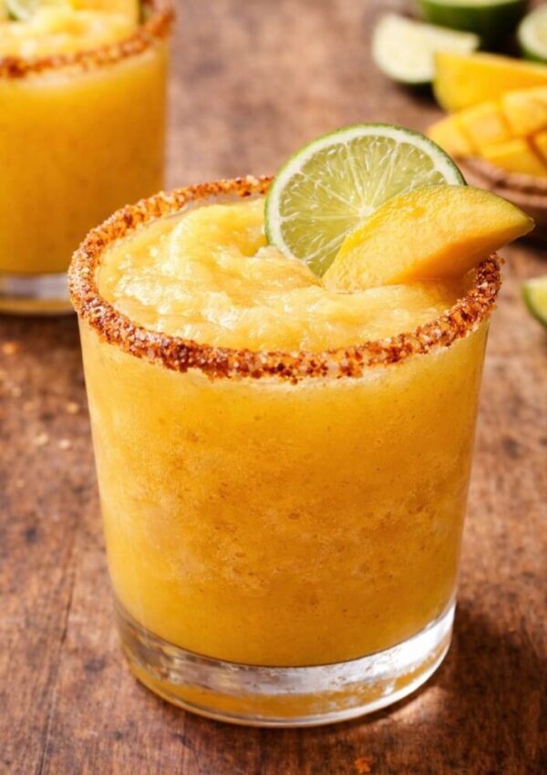 A glass of frozen mango margarita with a chili powder rim, garnished with a lime wheel and a mango slice, sits on a rustic wooden surface. Another glass and sliced fruit are visible in the background.