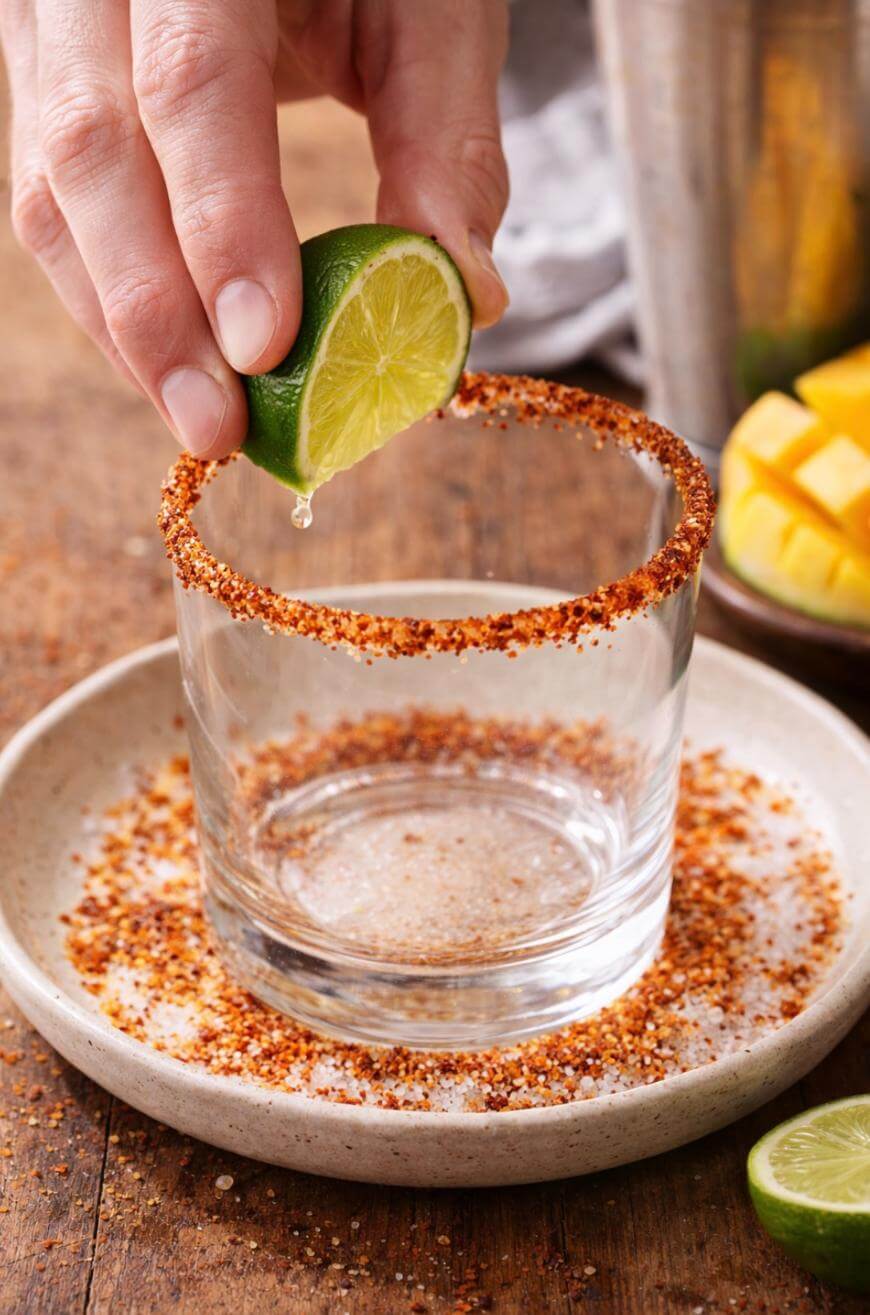 A hand squeezes a lime wedge over the rim of an empty glass coated with chili and salt, with a plate below catching the seasoning. A sliced lime and mango are visible in the background.