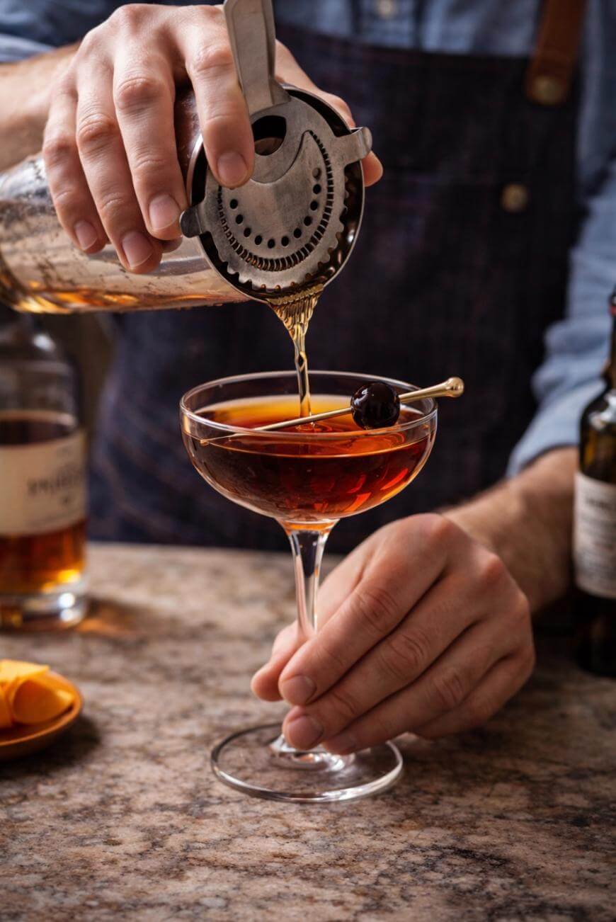 A bartender strains a dark cocktail into a coupe glass garnished with a cherry. The scene includes bottles and an orange peel on a marble countertop.