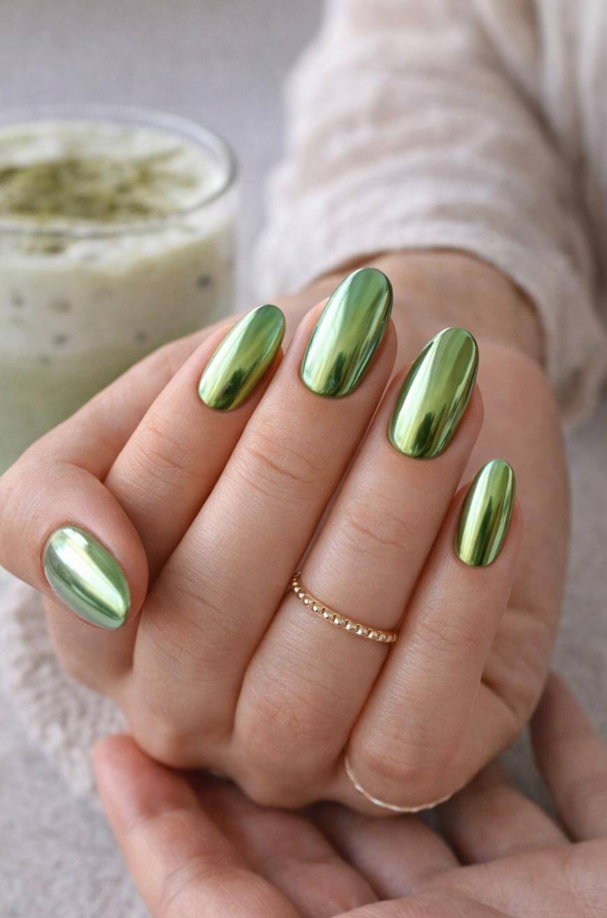 A hand with long, oval-shaped nails painted in shiny metallic green polish is shown close-up. The person wears a delicate gold ring and holds their hand gently. A green drink is blurred in the background.