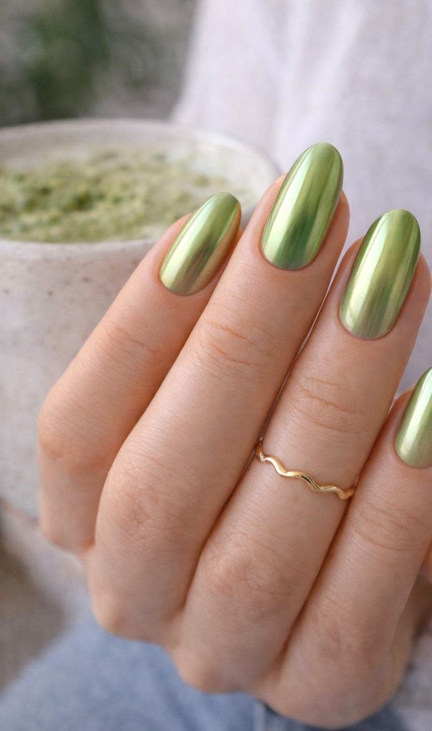 A hand with long, glossy, metallic green nails holds a speckled cup with a frothy green drink. The person wears a thin, wavy gold ring on their finger.