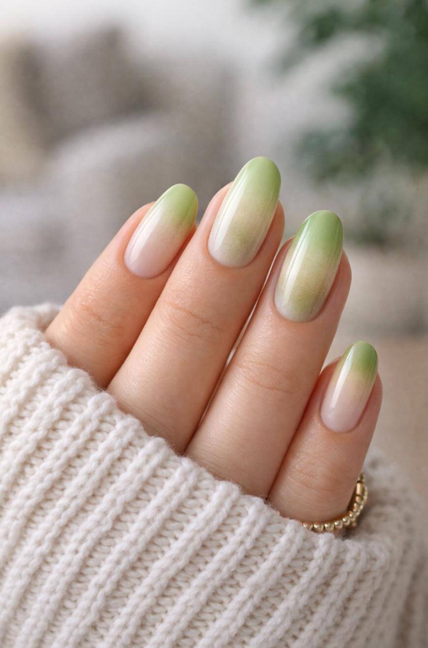 A hand with almond-shaped nails featuring a green and nude ombre design is shown, partially wrapped in a cozy, cream-colored knit sweater. The background is softly blurred.