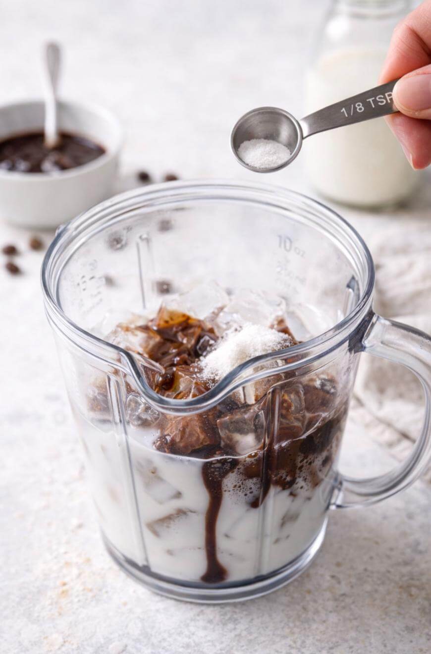 A hand adds a small spoonful of white powder to a blender filled with iced milk and coffee, with a jar and bowl of ingredients in the background.