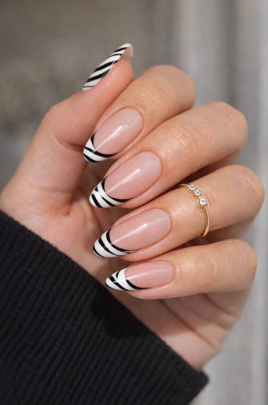 A hand with long almond-shaped nails featuring a nude base and black-and-white abstract striped tips. The person is wearing a gold ring with three small diamonds and a dark sleeve.