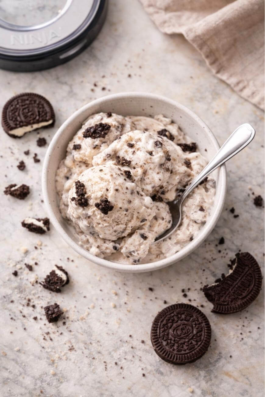 A bowl of cookies and cream ice cream with a spoon, surrounded by whole and crumbled chocolate sandwich cookies on a light marble surface.
