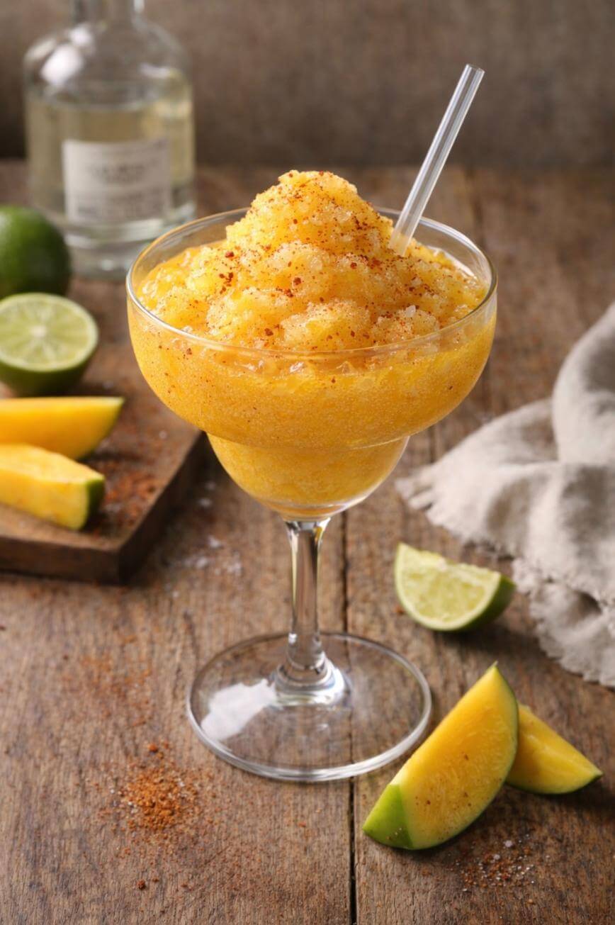 A frozen mango margarita topped with chili powder is served in a cocktail glass with a straw. Slices of mango, lime wedges, and a bottle are on a rustic wooden table in the background.