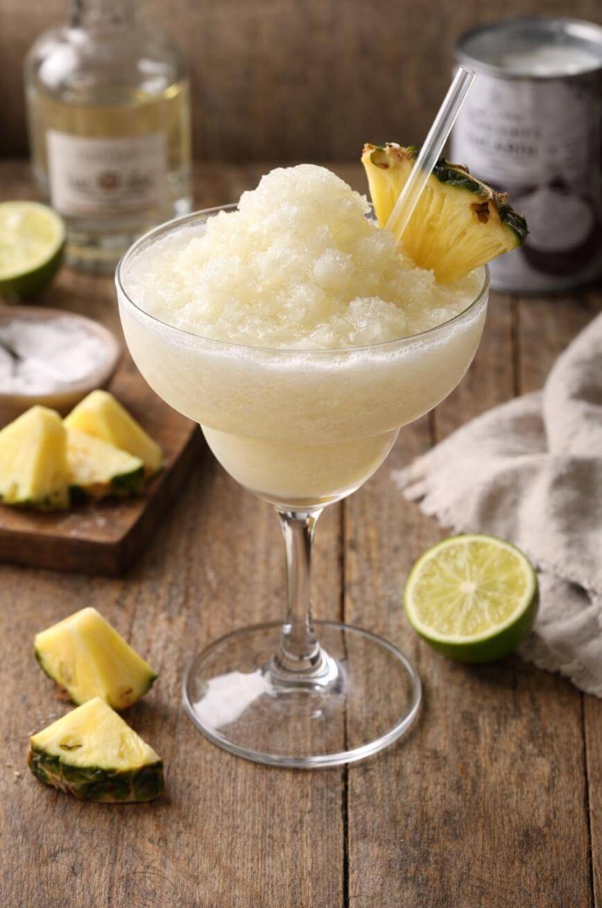 A frozen piña colada in a margarita glass, garnished with a pineapple slice and a straw, sits on a wooden table surrounded by fresh lime and pineapple pieces. A bottle and a can are blurred in the background.