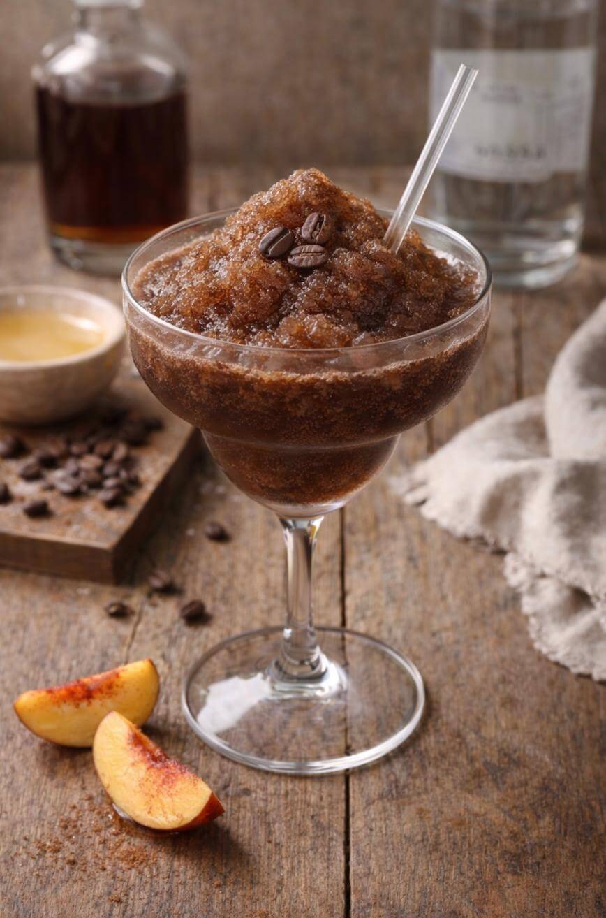 A glass filled with coffee granita topped with coffee beans and a straw sits on a rustic wooden table, surrounded by coffee beans, a bowl of syrup, peach slices, and bottles in the background.