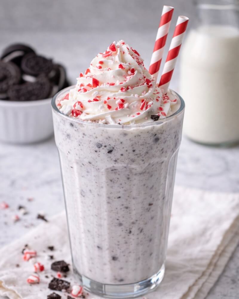 A cookies and cream milkshake topped with whipped cream and crushed peppermint, served in a glass with two red and white striped straws. A bowl of cookies and a bottle of milk are in the background.