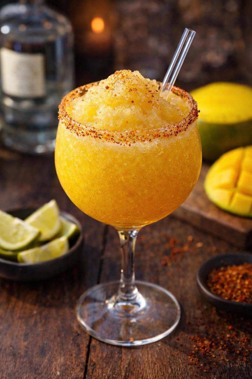 A frozen mango margarita in a stemmed glass with a chili powder rim and a straw, set on a rustic wooden table with lime wedges, a sliced mango, and a bottle in the background.